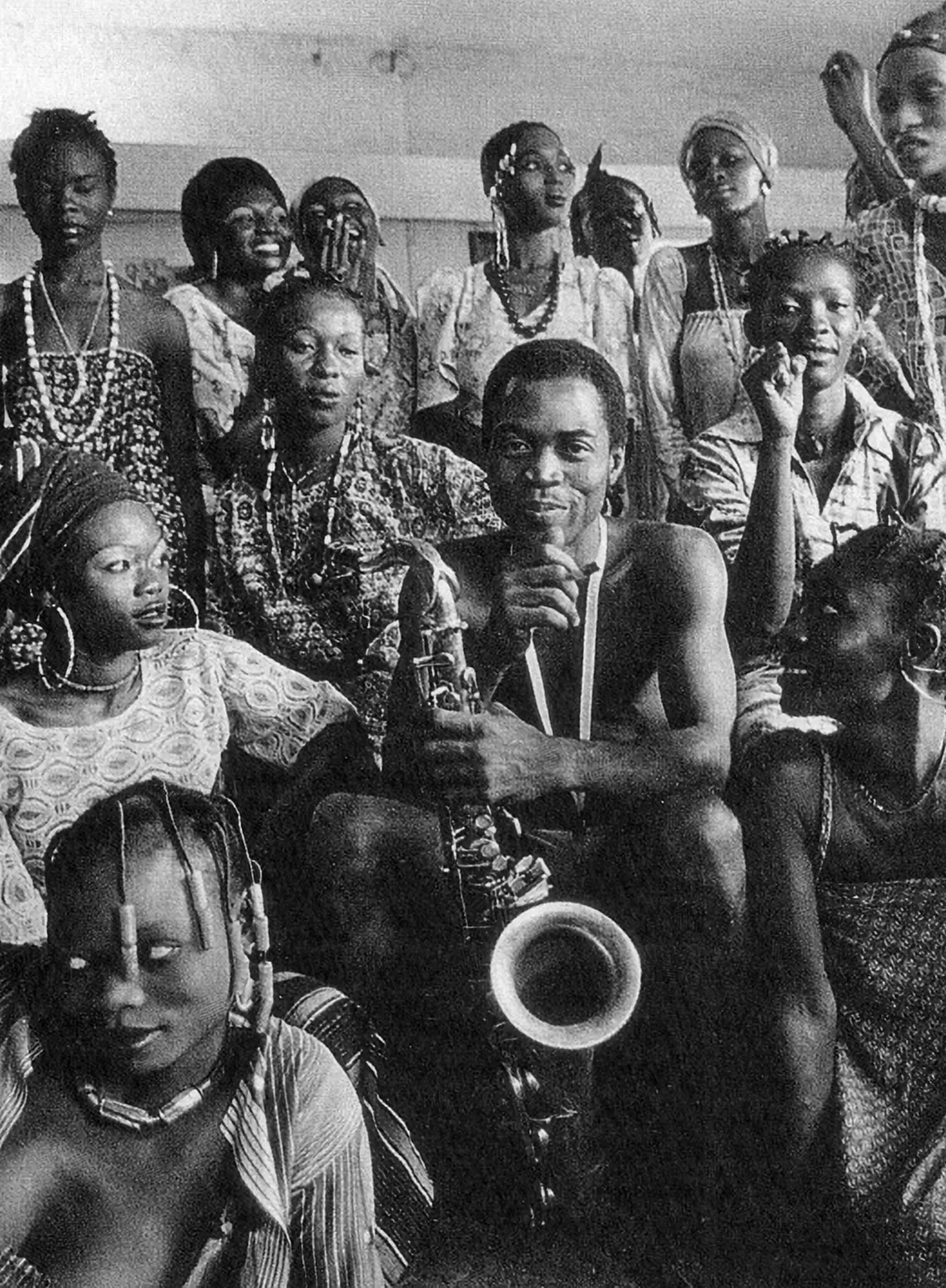 Fela Kuti sits smiling with his saxophone, surrounded on all sides by 17 of his wives.
